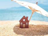 Miniature house and umbrella on beach, blue sea and sky on blurred background. Vacation home for happy holiday for family