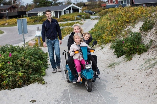 Familie mit Mutter im E-Rollstuhl macht Urlaub in einem Ferienhausgebiet in Dänemark