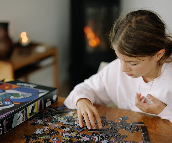 Ein Mädchen sitzt am Tisch und macht ein Puzzle. Im Hintergrund sorgt ein knisternder Holzofen für eine gemütliche Atmosphäre.